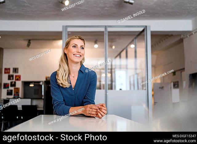Smiling female professional looking away while leaning on desk in office