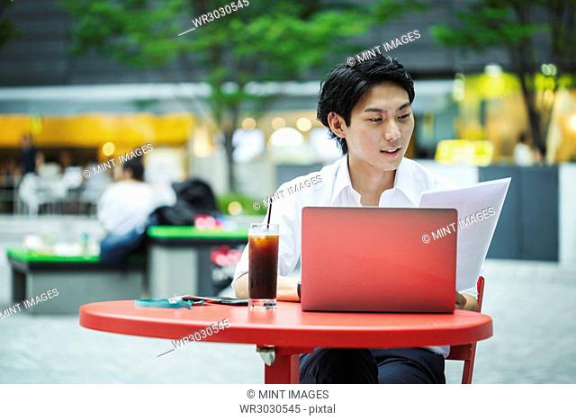 Businessman wearing white shirt sitting outdoors at red table, holding papers, working on laptop