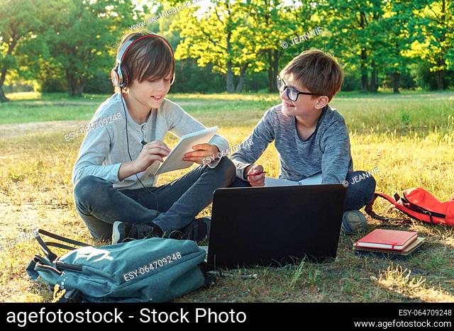 Loveable boy in glasses smiling happily while looking at pupil with earphones who reading school task from notebook. Two classmates enjoying studying together...