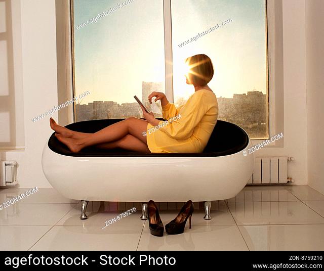 Adult woman in yellow dress relaxing on modern sofa with tablet. Sun shining through window