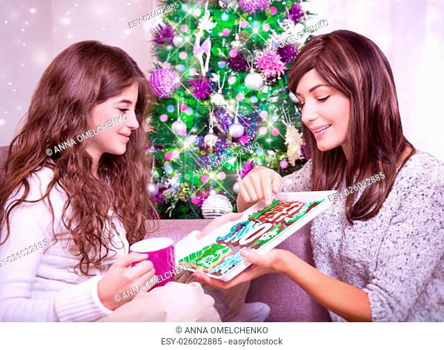 Portrait of cute mother with adorable teen daughter in Christmas eve at home near festive decorated fir tree, tasting beautiful traditional Xmas ginger cookies