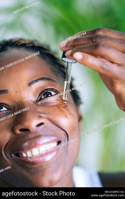 Afro woman smiling while applying face serum through dropper