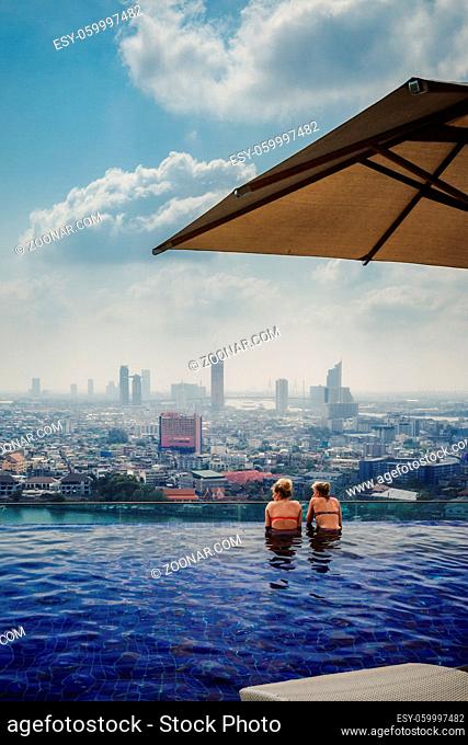 Two Girls in Bikinis Swimming in Infinity Pool Overlooking Bangkok Thailand Urban City Landscape Luxurious Vacation