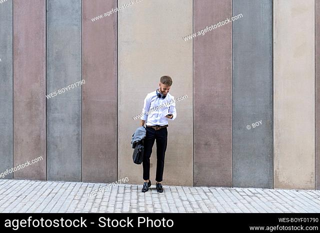 Mid adult businessman using mobile phone while standing with briefcase and jacket against wall