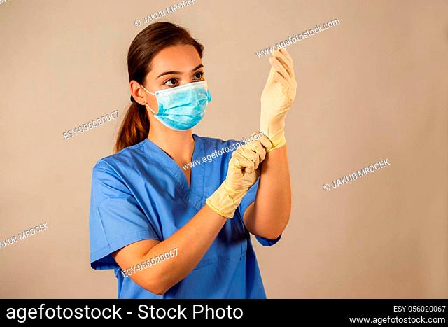 Adult woman looking at her hands with rubber gloves. Female nurse in blue shirt and face mask in studio. Concept of protection against viruses in hospital