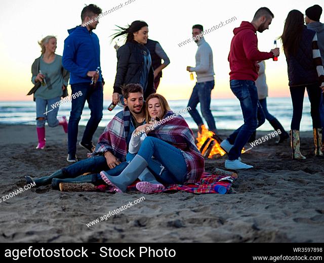 Young Couple Sitting with friends Around Campfire on The Beach At sunset drinking beer