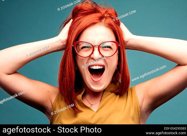 Front view of happy and positivity red haired woman in eyeglasses shouting holding head by hands, looking at camera. Emotionally girl smiling and surprised