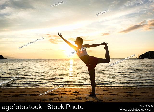 Full length side view of a fit woman practicing on the beach. The standing bow pulling pose for muscular strength and flexibility during summer vacation in...