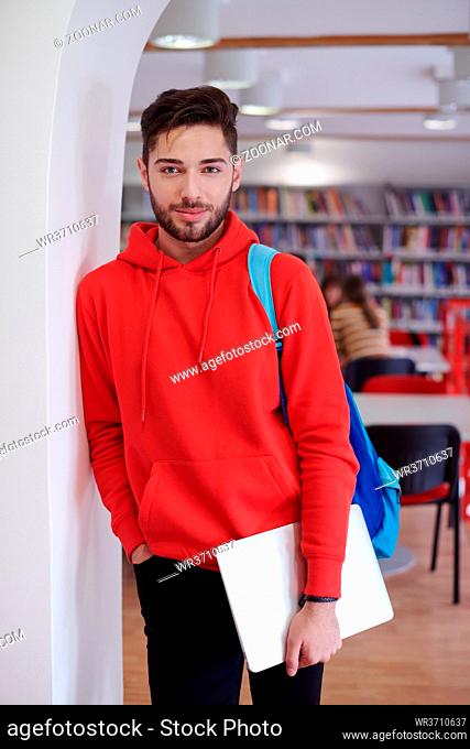 a young student is standing in front of the school library with a laptop in his hand