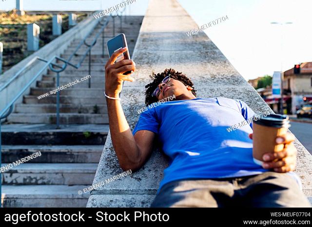 Young man with coffee using mobile phone lying on wall