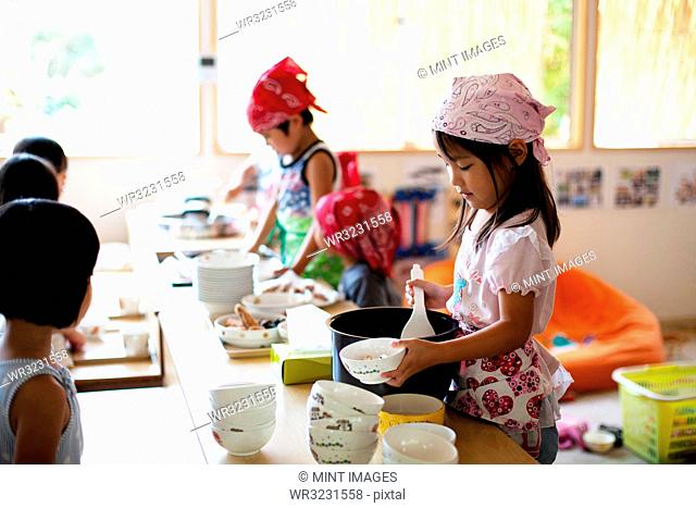 A girl and a boy wearing headscarves standing at a table in a Japanese preschool, serving lunch