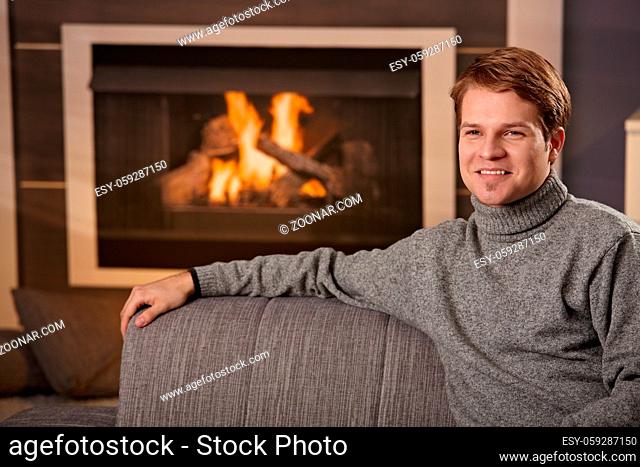 Young man sitting on sofa at home on a cold winter day in front of fireplace, smiling