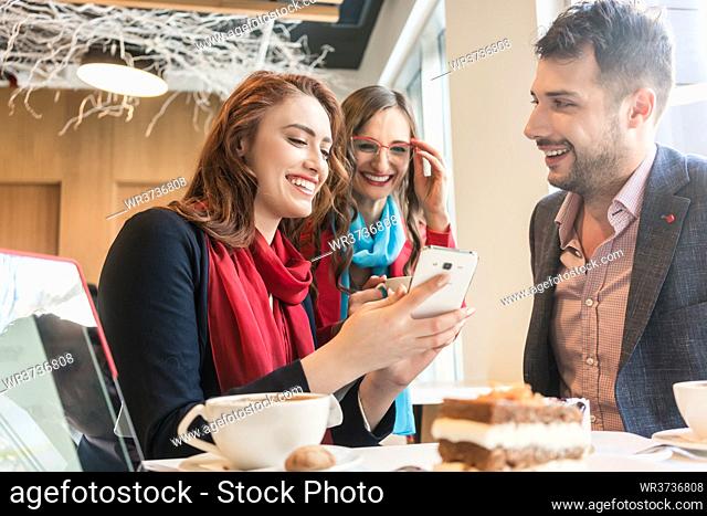 Three young colleagues or friends smiling while using a mobile phone for fun on social media during a coffee break in a trendy cafeteria