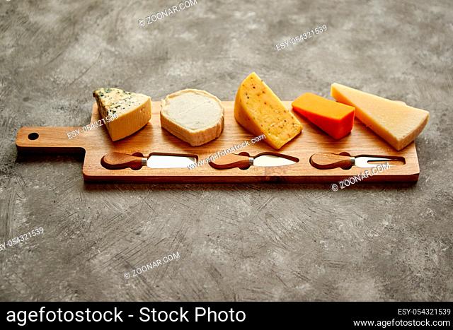 Assortment of various kinds of cheeses served on wooden board with fork and knives. Placed on concrete background