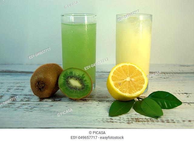 Two glass glasses with a refreshing juice and ice, on a textile stand, whole and sliced half of an orange with leaves and kiwi, lies on a white wooden table