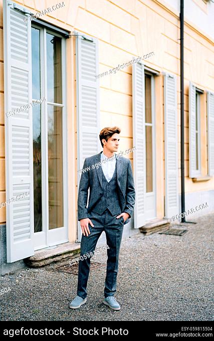 Serious groom looks into the distance and stands at the door of an old shuttered house. Lake Como, Italy. High quality photo