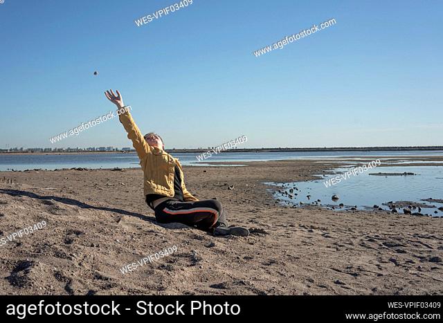 Boy sitting alone on sandy beach and throwing stones