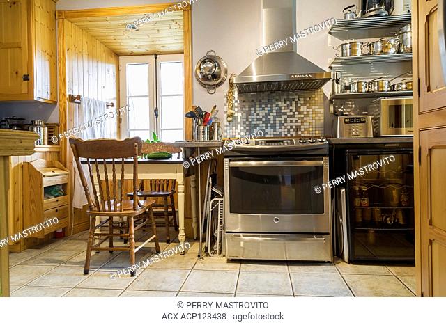 Stainless steel oven and white antique wooden breakfast table and chairs, pinewood cabinets in kitchen with ceramic tile floor inside an old 1820 cottage style...