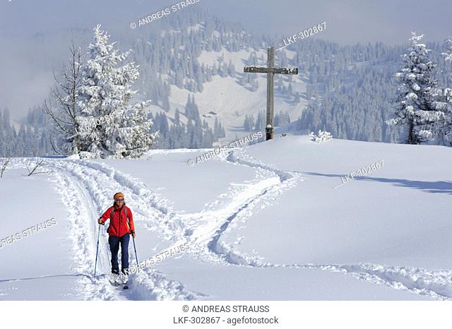 Woman backcountry skiing ascending in a track in front of summit cross and alpine huts, Hochries, Chiemgau range, Upper Bavaria, Bavaria, Germany, Europe