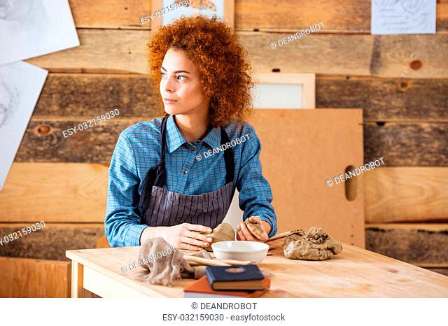 Thoughtful curly young woman sitting in pottery workshop and making sculpture with clay