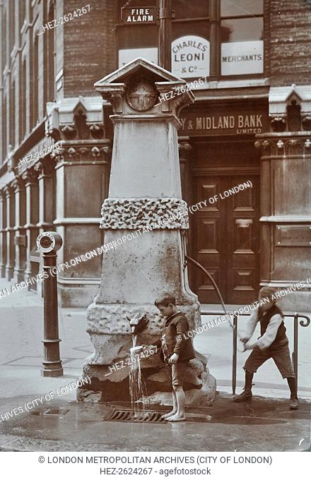 Children drawing water from the Aldgate pump, London, August 1908. One boy pumps while the other holds a cup under the water outlet