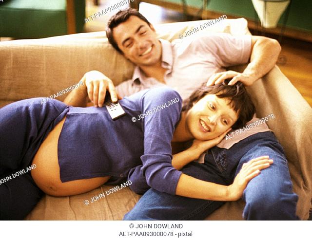 Man and pregnant woman on sofa, woman lying on man's lap, man holding remote control, portrait