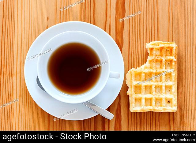Wooden table with white cup with saucer and spoon as well as lying waffle with bite mark in top view