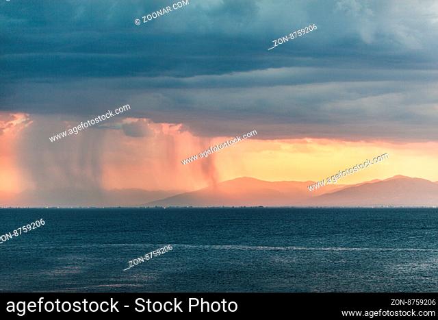 Rain pouring from clouds over distant plains near mountains with orange sun light at the horizon