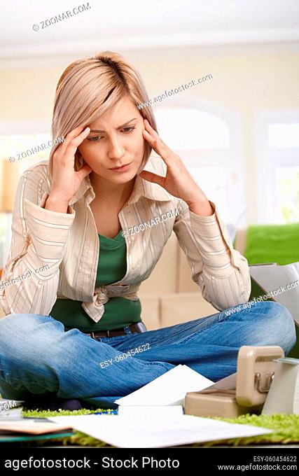 Troubled woman holding head sitting on living room floor looking at documents