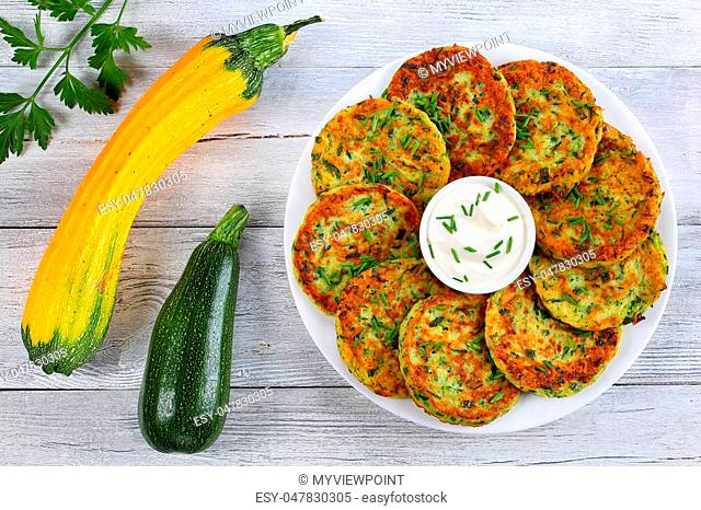 delicious zucchini fritters on white plate with sour cream in bowl in center of platter sprinkled with finely chopped chives, raw zucchini on wooden table
