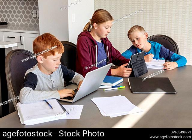 A teenage girl and young boys sit at the kitchen table at home with a laptop and tablet doing school work while being homeschooled; Edmonton, Alberta, Canada