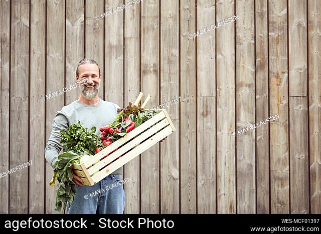 Smiling mature man carrying vegetable crate while standing against house