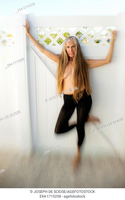 Partially nude 57 year old woman with long hair smiling at the camera standing on one leg against a gate