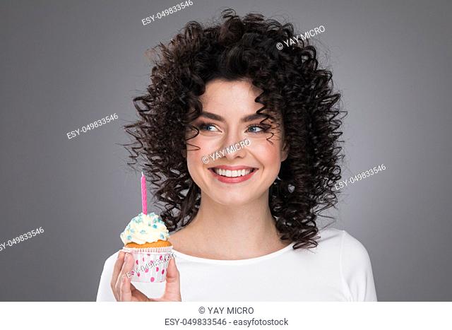 Cheerful beautiful curly young woman making a wish holding birthday cupcake with candle