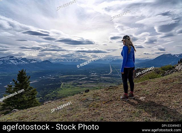 View from behind of a woman standing on a mountaintop looking out at the vista of the majestic mountain ranges and valley below with a dramatic cloudy sky on a...