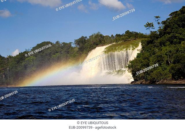 Canaima Village waterfalls feeding into Canaima lake while the humidity of its waters are producing a rainbow effect with blue sky and white clouds