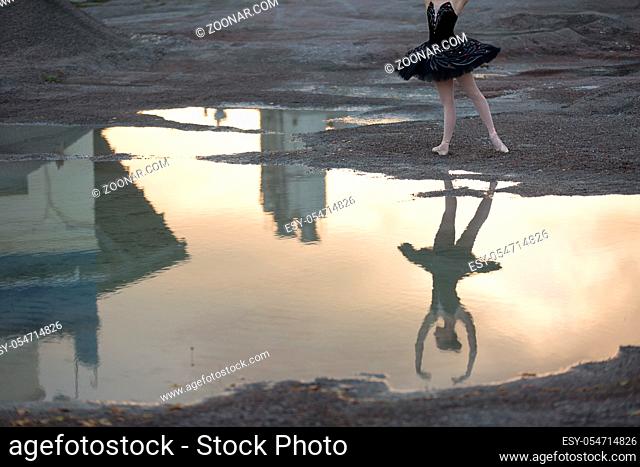 Nice reflection on the water of a young ballerina in a black tutu against the sky and technical structures. She stands on the gravel. Outdoor
