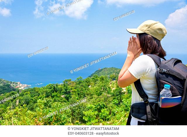 Women tourist with a backpack wear cap raise hand at the mouth yelling and beautiful nature landscape blue sea and sky from high scenic viewpoint at Koh Tao
