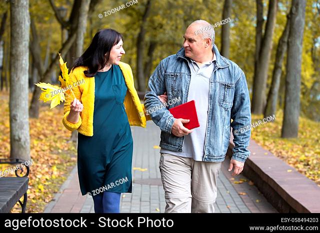 A beautiful middle-aged couple is walking in the autumn park. An elderly man and woman are walking along the road