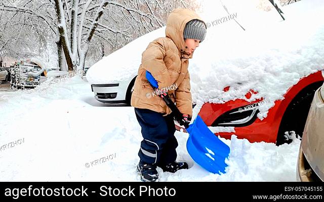 Smiling boy in jacket and hat helping to clean up the snow covered car after blizzard using big blue shovel