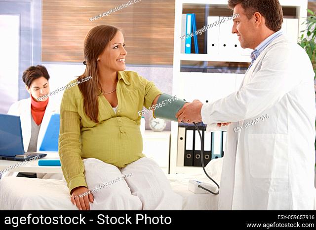 Expectant woman sitting in consulting room, doctor checking blood pressure, assistant at desk with laptop in background