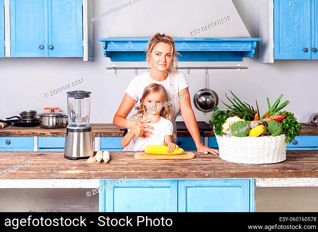 Happy attractive mother hugging daughter, standing together in kitchen with modern furniture, little girl holding corn, basket of fresh vegetables on table
