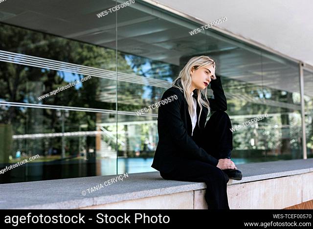 Pensive young businesswoman with dyed blond hair sitting in front of office building looking at distance
