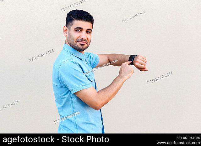 Portrait of satisfied handsome young bearded man in blue shirt standing, pointing and holding his smart watch, looking at camera with smile