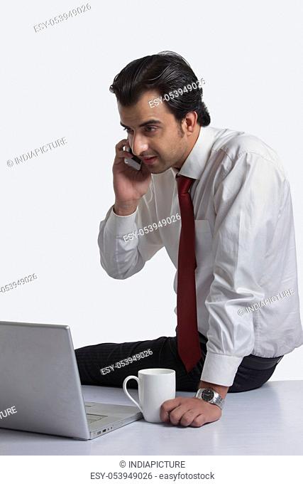 Young businessman answering mobile phone while looking at laptop over white background