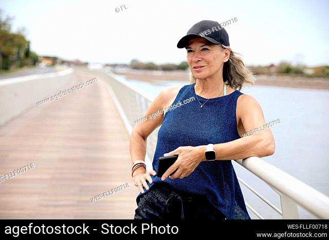 Thoughtful senior woman holding mobile phone standing by railing