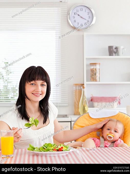 Pretty brunette woman eating a salad next to her baby while sitt