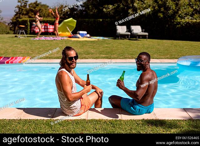 Two diverse male friends drinking beer and sitting at the poolside