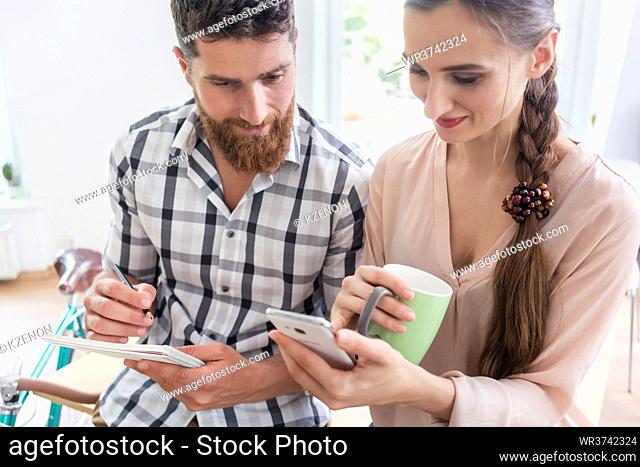 Two young co-workers smiling while looking at a funny photo or watching a video on the mobile phone, during break in a modern shared office space