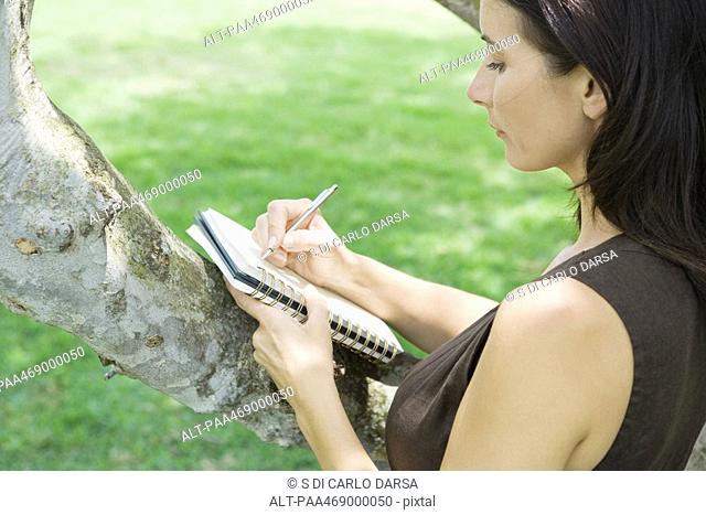 Woman writing in notebook, holding notebook against branch of tree, high angle view